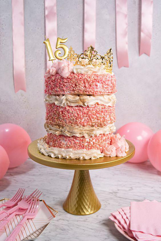 A pink, three-tiered quinceañera ice cream cake sits upon a golden cake stand. The cake is decorated with white and pink icing, and it has two candles that make up the number “15.” Beside the candles rests a tiara. In the foreground, to the right, there’s a stack of pink napkins on top of pink disposable plates. In the foreground to the left, there’s a stack of mid-century modern-esque plates with pink disposable forks on top. In the background on the wall are pink, vertical streamers. Pink balloons sit in the background to the left and right sides of the cake.