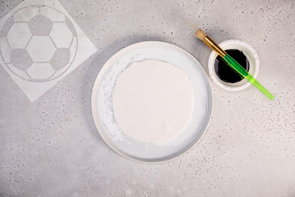 A stencil of a soccer ball pattern rests in the top left corner. In the center, there’s a plate lined with powdered sugar and a circular cutout of fondant resting on top. Next to the plate on the right is a small cup/bowl with black food coloring and a small paintbrush sitting on top.