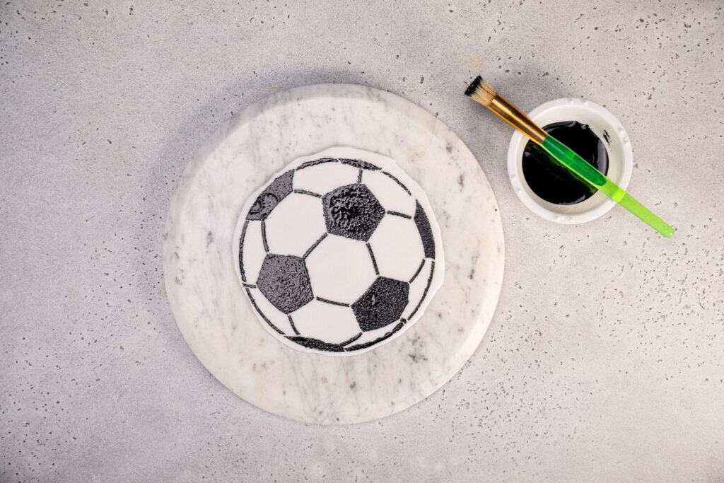 The finished soccer ball pattern that’s been painted onto the fondant rests on top of the stone plate, and a paintbrush sits on top of the bowl of black food coloring.
