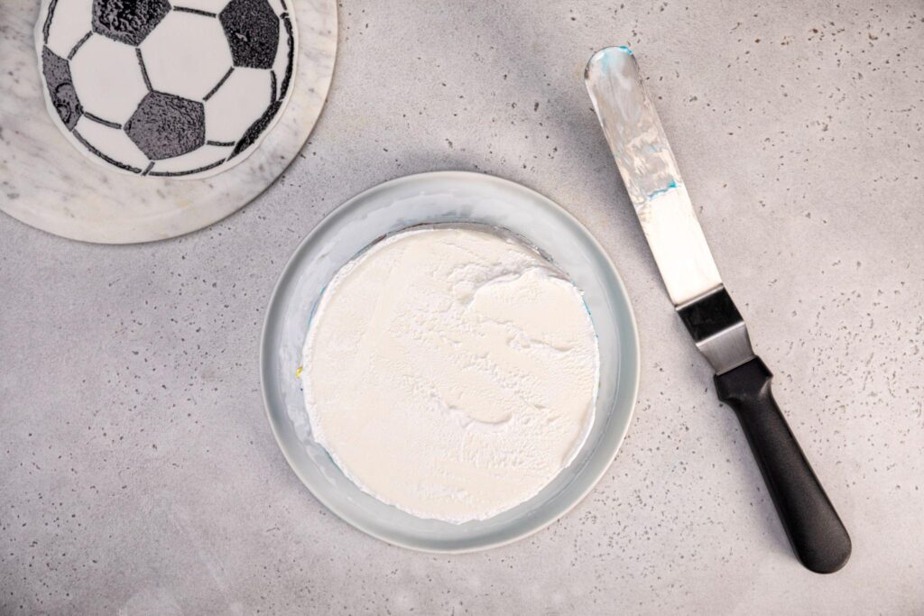 The fondant with the soccer ball pattern remains in the top left corner of the image. A Carvel Happy Birthday Ice Cream Cake without any icing on the top or sides sits in the middle of the image, while the icing knife rests next to the cake. 