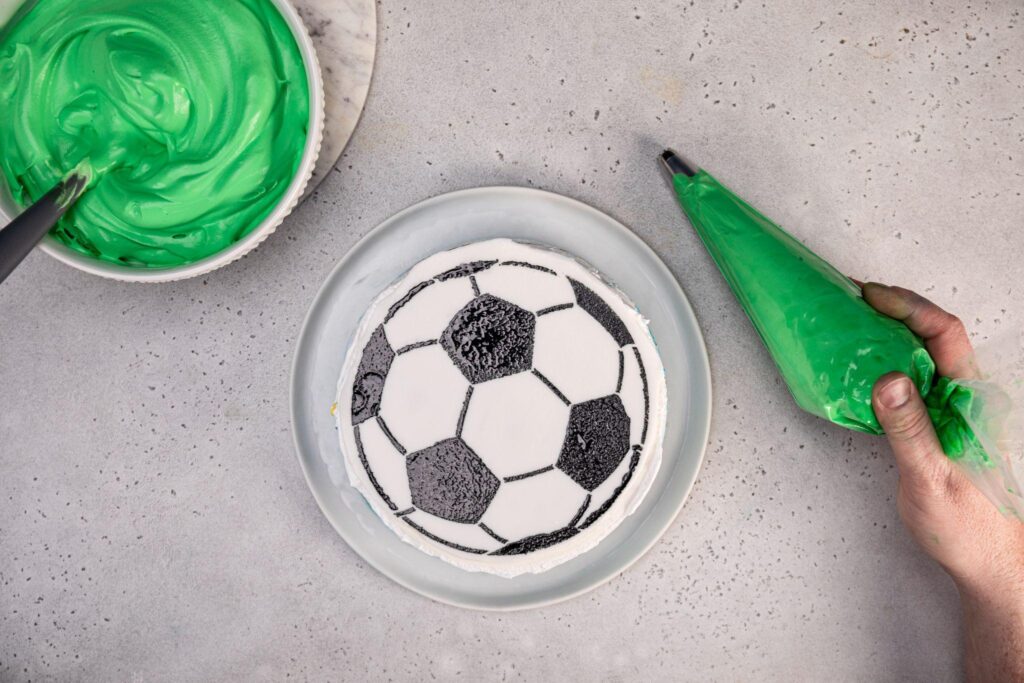 A large bowl of green icing with a spoon in it rests on the stone plate in the top left corner. The soccer ball cake is in the middle, and a hand on the right holds an icing piping bag that’s full of green icing. It has a grass tip attached to the piping end.